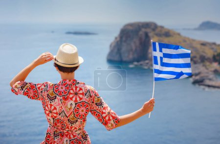 Foto de Agradable feliz hembra disfrutando del día soleado en las islas griegas. Viaje a Grecia, islas mediterráneas fuera de temporada turística. Mujer viajera joven con bandera griega nacional disfrutando de la vista al mar - Imagen libre de derechos