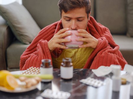 Young Caucasian man with flu on sofa at home. Young Caucasian man with flu feels cold in living room at home drink hot tea in cup. in front of him table are traditional and alternative medicine