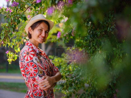 young asian woman stands in sunny green park, admiring blooming bougainvillea bush. The flowers bright colors are beautiful contrast to parks lush greenery.