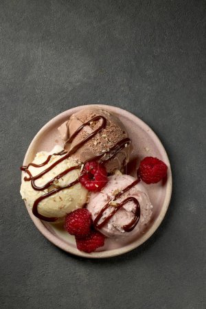 Foto de Plate of various ice cream balls decorated with chocolate sauce and fresh rasberries on dark grey background, top view - Imagen libre de derechos