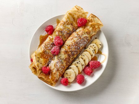 Photo for Freshly baked crepes decorated with raspberries, banana and caramel sauce on white plate, top view - Royalty Free Image