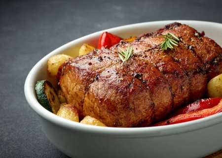 Photo for Bowl of whole roast pork and vegetables on dark grey kitchen table - Royalty Free Image