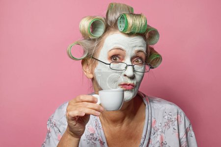 Foto de Mujer mayor divertida con mascarilla cosmética y taza de café sobre fondo rosa - Imagen libre de derechos