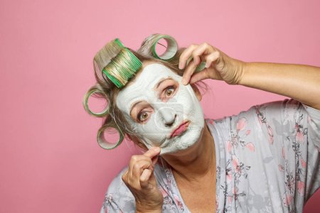Foto de Mujeres mayores divertidas con mascarilla de arcilla y rollos de pelo sobre fondo rosa - Imagen libre de derechos