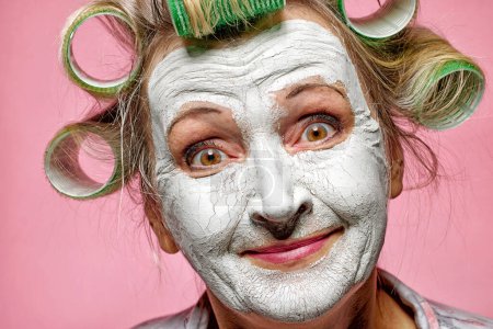 Foto de Mujer mayor divertida con mascarilla de arcilla y rollos de pelo sobre fondo rosa - Imagen libre de derechos