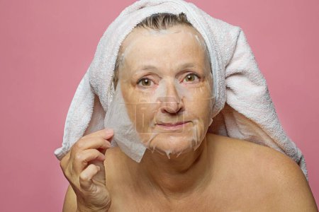 Téléchargez les photos : Senior femme appliquant humide masque facial serviette sur fond rose - en image libre de droit