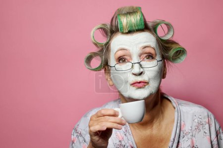Téléchargez les photos : Drôle de femme âgée avec masque en argile et rouleaux de cheveux prendre un café le matin - en image libre de droit
