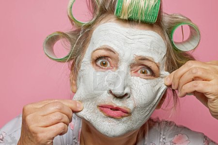 Foto de Retrato de mujer mayor divertida con mascarilla de arcilla y rodillos de pelo que quiere cambiar su piel y lucir más joven - Imagen libre de derechos