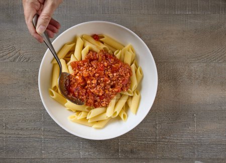 Téléchargez les photos : Sauce bolognaise est ajoutée à un bol de pâtes penne, vue du dessus - en image libre de droit