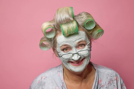 Foto de Retrato de mujer mayor sonriente divertida con rodillos de pelo, mascarilla de arcilla y gafas sobre fondo rosa - Imagen libre de derechos