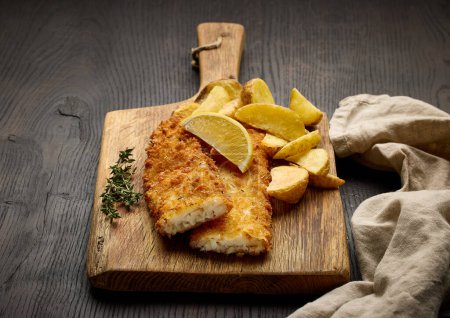 Téléchargez les photos : Filets de poisson panés et quartiers de pommes de terre frites servis sur planche à découper en bois - en image libre de droit