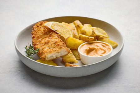 Téléchargez les photos : Un bol de fish and chips. Filet de poisson pané avec quartiers de pommes de terre frites sur table de cuisine gris clair - en image libre de droit