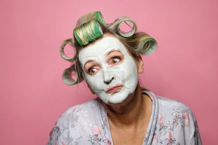 Téléchargez les photos : Drôle réfléchie femme âgée avec des rouleaux de cheveux et masque d'argile sur fond rose - en image libre de droit