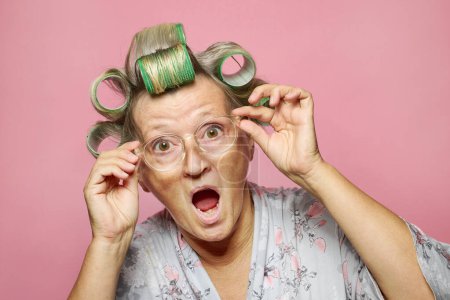 Foto de Divertido sorprendido sorprendió a las mujeres mayores con rodillos de pelo y gafas sobre fondo rosa - Imagen libre de derechos