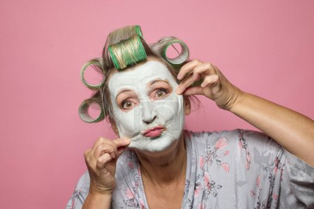 Foto de Mujeres mayores divertidas con mascarilla de arcilla y rollos de pelo sobre fondo rosa - Imagen libre de derechos