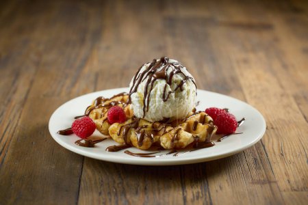 Photo for Plate of belgian waffle with vanilla ice cream, melted chocolate sauce and raspberries on wooden table - Royalty Free Image