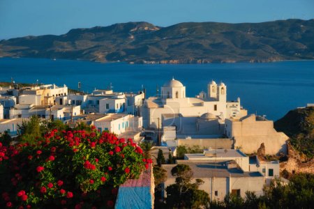 Blick auf das Dorf Plaka auf der Insel Milos über rote Geranienblüten bei Sonnenuntergang. Plaka Stadt, Insel Milos, Griechenland.. Fokus auf Blumen