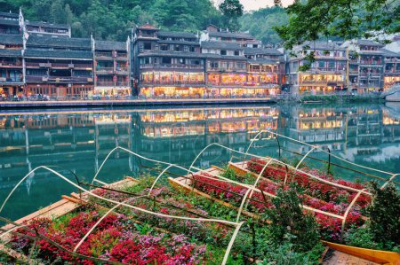 Photo for Chinese tourist attraction destination - Feng Huang Ancient Town (Phoenix Ancient Town) on Tuo Jiang River with flower beds on boats illuminated at night. Hunan Province, China - Royalty Free Image