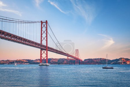 Photo for View of 25 de Abril Bridge famous tourist landmark over Tagus river, Christ the King monument and a tourist yacht boat at sunset. Lisbon, Portugal - Royalty Free Image