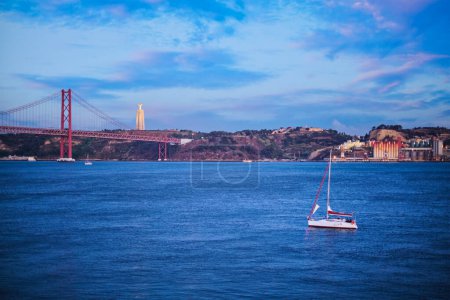 Photo for View of 25 de Abril Bridge over Tagus river, Christ the King monument and a yacht boat in the evening twilight. Lisbon, Portugal - Royalty Free Image