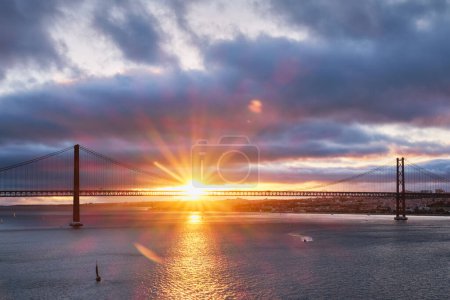 Photo for View of 25 de Abril Bridge famous tourist landmark of Lisbon connecting Lisboa and Almada on Setubal Peninsula over Tagus river with tourist yacht boats at sunset. Lisbon, Portugal - Royalty Free Image
