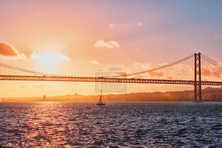 Photo for View of 25 de Abril Bridge famous tourist landmark of Lisbon connecting Lisboa and Almada on Setubal Peninsula over Tagus river with tourist yacht silhouette at sunset. Lisbon, Portugal - Royalty Free Image