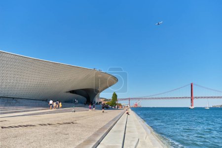 Foto de Lisboa, Portugal - 08 de agosto de 2022: MAAT Museo de Arte, Arquitectura y Tecnología es un edificio moderno a orillas del río Tajo y 25 de abril puente en el fondo con avión volador. Lisboa, Portugal - Imagen libre de derechos