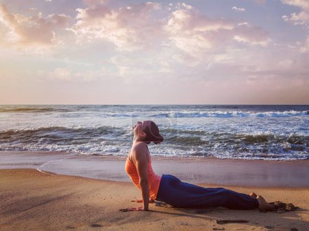 Foto de Yoga al aire libre - mujer haciendo Ashtanga Vinyasa yoga Surya Namaskar Sun Salutation asana Urdhva Mukha Svanasana - postura hacia arriba del perro en la puesta de sol en la playa. Vintage efecto retro imagen hipster filtrado - Imagen libre de derechos