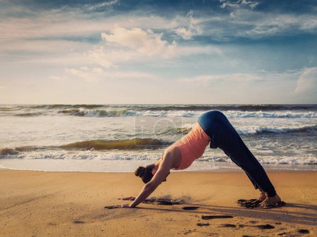 Foto de Imagen hipster filtrada efecto retro vintage de Yoga al aire libre - mujer haciendo Ashtanga Vinyasa yoga asana Adho mukha svanasana - perro orientado hacia abajo - en la playa tropical al atardecer. Kerala, India - Imagen libre de derechos