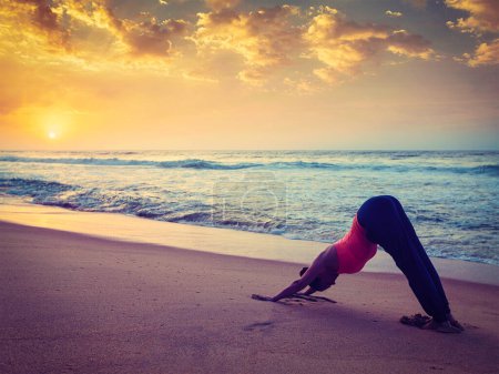 Téléchargez les photos : Vintage effet rétro filtré hipster style image de Yoga en plein air femme faisant Ashtanga Vinyasa yoga asana Adho mukha svanasana - chien orienté vers le bas - à la plage tropicale au coucher du soleil. Kerala, Inde - en image libre de droit