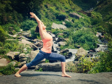 Téléchargez les photos : Yoga en plein air - femme en forme sportive faisant Ashtanga Vinyasa Yoga asana Virabhadrasana 1 posture de guerrier à la cascade dans les montagnes HImalayas. Vintage effet rétro filtré image de style hipster
. - en image libre de droit