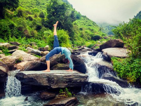 Photo for Yoga outdooors - woman doing yoga asana eka pada urdva dhanurasana Upward Bow Pose back benkd outdoors at waterfall in Himalayas. Vintage retro effect filtered hipster style image. - Royalty Free Image