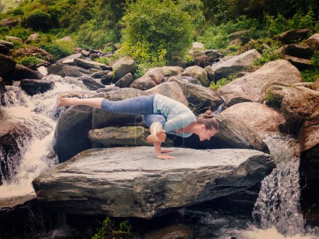 Foto de Joven mujer en forma deportiva haciendo yoga asana Eka Pada Koundinyasana 1 en la cascada tropical. Vintage efecto retro filtrado hipster estilo imagen. - Imagen libre de derechos