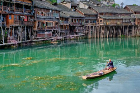 Foto de FENGHUANG, CHINA - 23 DE ABRIL DE 2018: Hombre chino no identificado en el barco Feng Huang Ancient Town (Phoenix Ancient Town) en el río Tuo Jiang con puente y barco turístico. Provincia de Hunan, China - Imagen libre de derechos