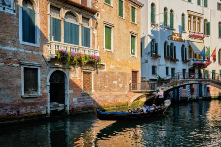 Foto de VENECIA, ITALIA - 19 DE JULIO DE 2019: Estrecho canal entre coloridas casas antiguas con góndola barco con turistas y gonolier en Venecia, Italia - Imagen libre de derechos