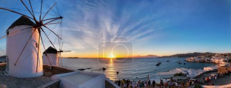 Photo for MYKONOS, GREECE - MAY 29, 2019: Panorama of traditional greek windmills on Mykonos island on sunset with dramatic sky and Little Venice quarter with tourist crowd - Royalty Free Image