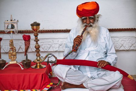 Photo for JODHPUR, INDIA - NOVEMBER 13, 2019: Old Indian man with beard smokes hookah (waterpipe) in Mehrangarh fort. The concept of hookah is thought to have originated in India - Royalty Free Image