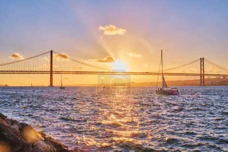 Photo for View of 25 de Abril Bridge famous tourist landmark of Lisbon connecting Lisboa and Almada on Setubal Peninsula over Tagus river with tourist yacht silhouette at sunset. Lisbon, Portugal - Royalty Free Image