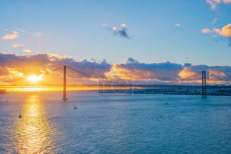 Photo for View of 25 de Abril Bridge famous tourist landmark of Lisbon connects Lisboa and Almada on Setubal Peninsula over Tagus river with tourist yacht silhouette at sunset and flying plane. Lisbon, Portugal - Royalty Free Image