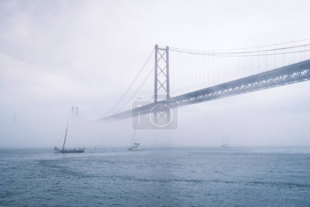 Foto de Vista del puente 25 de Abril famoso monumento turístico de Lisboa que conecta Lisboa y Almada en la niebla pesada niebla wtih barcos de yate que pasan por debajo. Lisboa, Portugal - Imagen libre de derechos