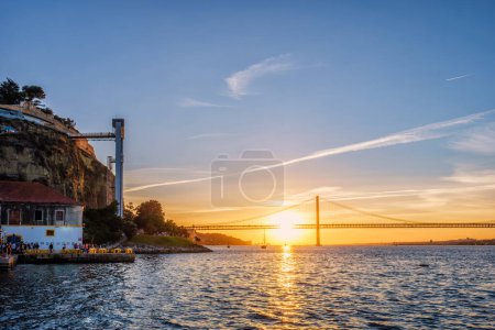 Photo for View of 25 de Abril Bridge famous tourist landmark of Lisbon over Tagus river and Boca do Vento Elevator and seaside restaurants with people at the sunset. Lisbon, Portugal - Royalty Free Image