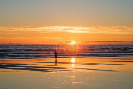 Foto de Puesta de sol del océano Atlántico con silueta de fotógrafo tomando imágenes de olas crecientes en la playa de Fonte da Telha, Costa da Caparica, Portugal - Imagen libre de derechos