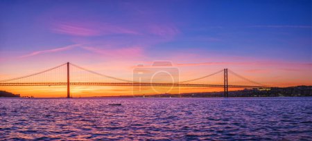 Photo for Panorama of 25 de Abril Bridge famous tourist landmark of Lisbon connecting Lisboa and Almada over Tagus river with tourist yacht silhouette at sunset. Lisbon, Portugal - Royalty Free Image
