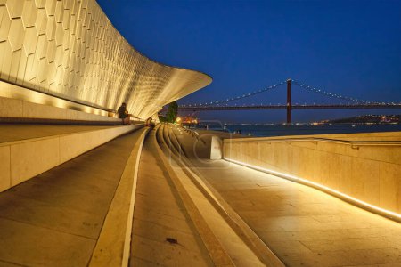 Foto de Lisboa, Portugal - 08 de agosto de 2022: MAAT - Museo de Arte, Arquitectura y Tecnología es un edificio moderno a orillas del río Tajo con el puente 25 de abril en el fondo por la noche. Lisboa, Portugal - Imagen libre de derechos