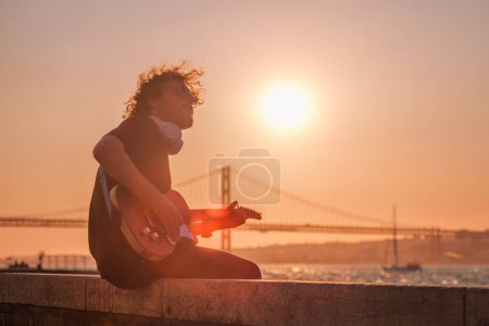Foto de Hipster músico callejero de negro tocando la guitarra eléctrica en la calle al atardecer en terraplén con 25 de abril puente en el fondo. Lisboa, Portugal - Imagen libre de derechos