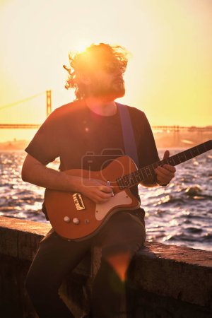 Téléchargez les photos : Musicien de rue Hipster en noir jouant de la guitare électrique dans la rue au coucher du soleil sur un remblai avec pont du 25 avril en arrière-plan. Lisbonne, Portugal - en image libre de droit