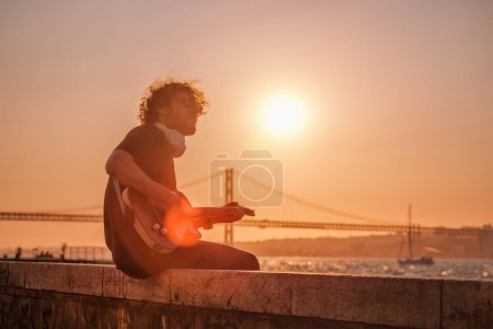 Téléchargez les photos : Musicien de rue Hipster en noir jouant de la guitare électrique dans la rue au coucher du soleil - en image libre de droit