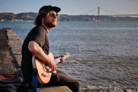 Téléchargez les photos : Musicien de rue Hipster en noir jouant de la guitare électrique dans la rue assis sur un quai au coucher du soleil à Lisbonne, Portugal - en image libre de droit