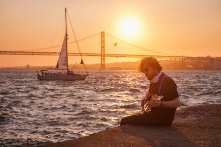 Téléchargez les photos : Musicien de rue Hipster en noir jouant de la guitare électrique dans la rue au coucher du soleil sur un remblai avec pont du 25 avril et yacht en arrière-plan. Lisbonne, Portugal - en image libre de droit