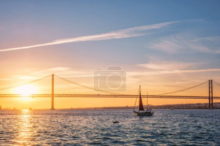 Photo for View of 25 de Abril Bridge famous tourist landmark of Lisbon connecting Lisboa and Almada over Tagus river with tourist yacht silhouette at sunset. Lisbon, Portugal - Royalty Free Image